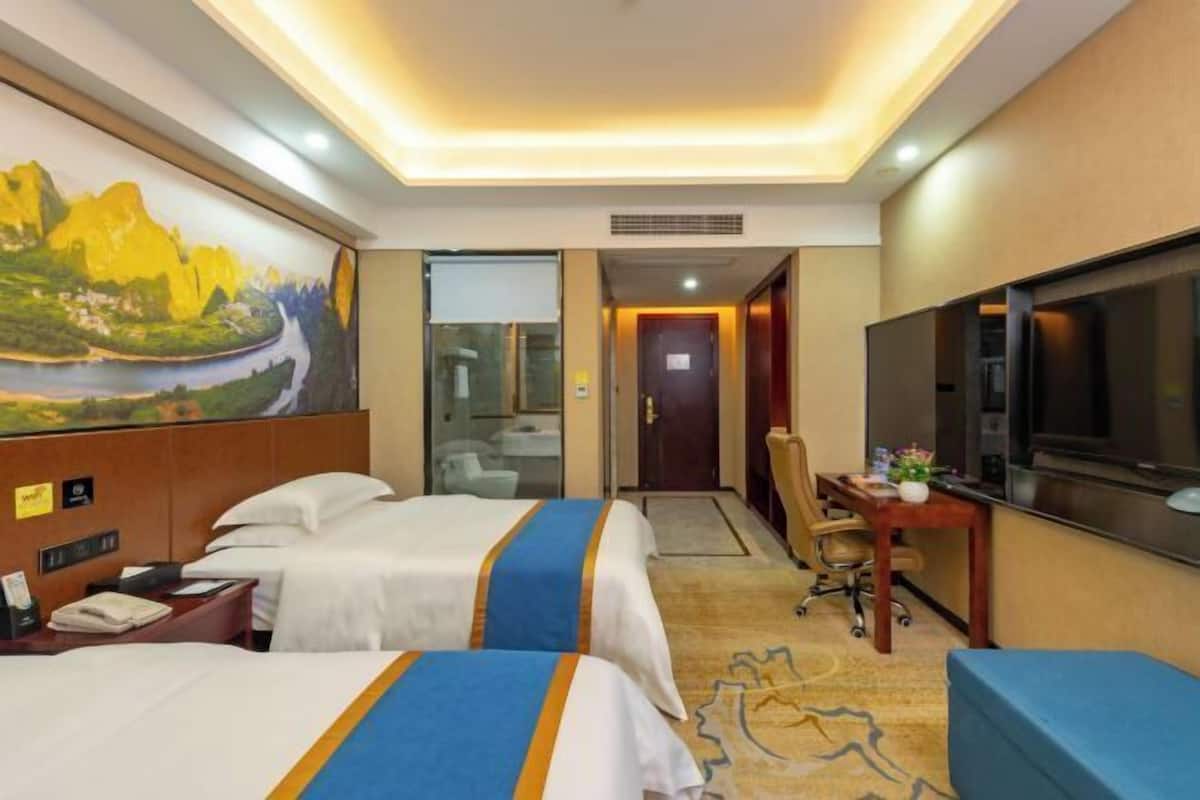 A hotel room with two beds, a desk, a television, and a painting on the wall.
