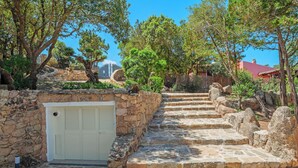 Una escalera de piedra conduce a un edificio con una puerta blanca, rodeado de árboles y un muro de piedra.