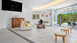 Un salón amplio con televisor de pantalla plana, un mueble de madera, un sofá con un cojín amarillo, una mesa de centro y una zona de comedor con mobiliario de exterior.