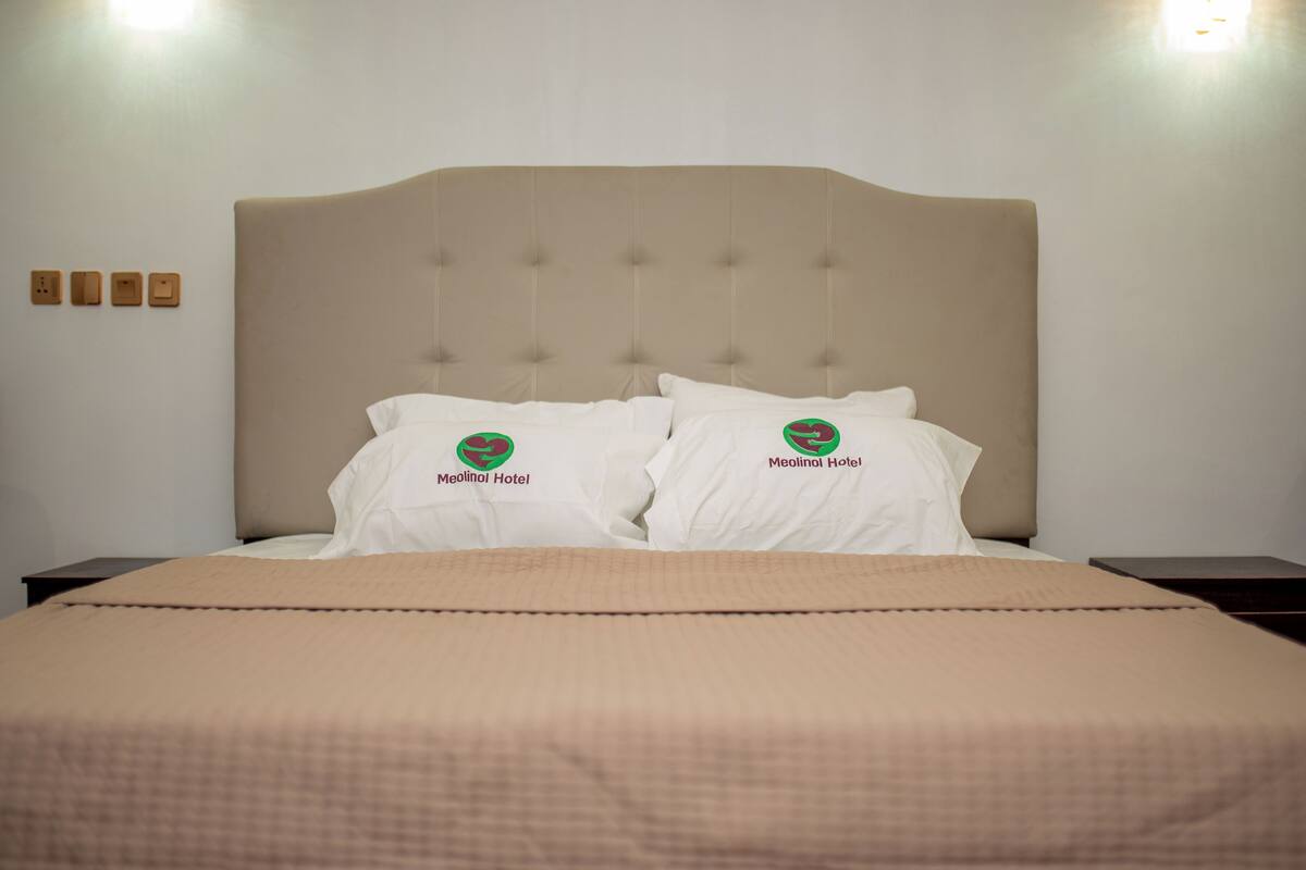 Una cama con cabecera color beige, dos almohadas blancas con un logotipo y una colcha color beige.