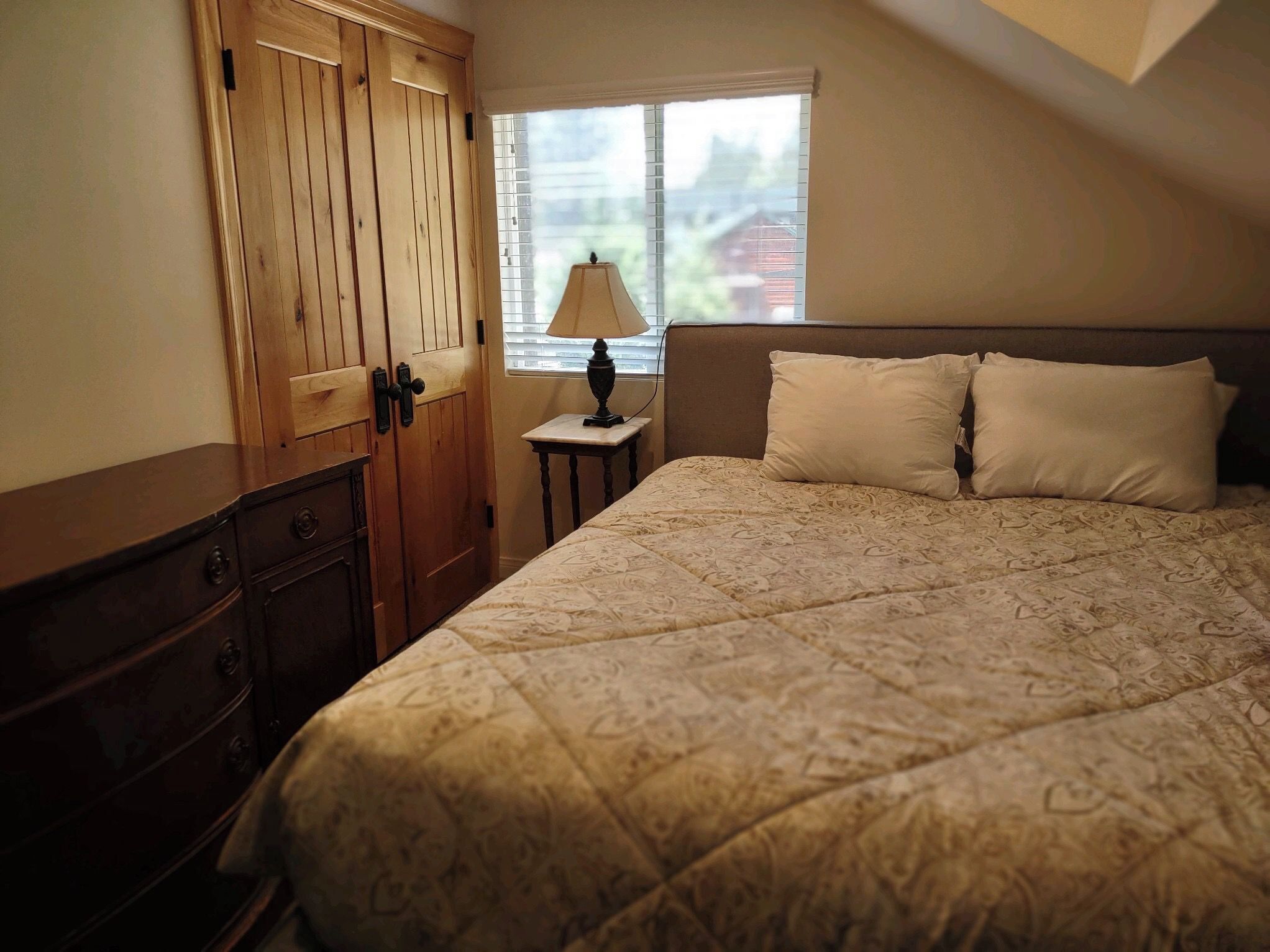 Een slaapkamer met een bed, nachtkastje, lamp, houten garderobe en een raam met jaloezieën.