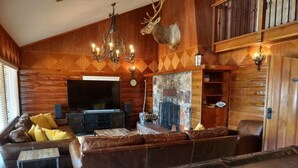 Interior de una cabaña de troncos con un sofá grande, un televisor, una chimenea y una cabeza de venado montada.