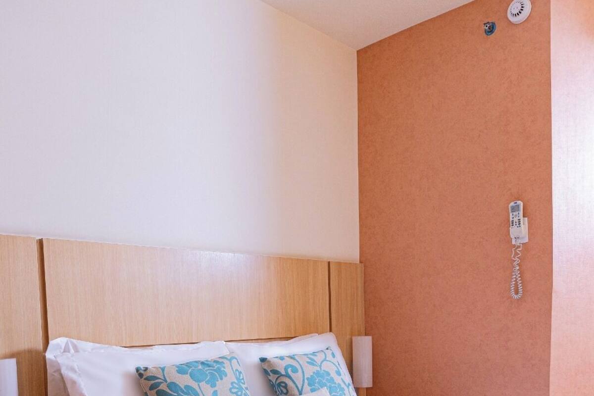 Ein ordentlich bezogenes Bett mit weiß-blauen blumigen Kissen, einem Holzkopfschild und einem an der Wand befestigten Telefon.