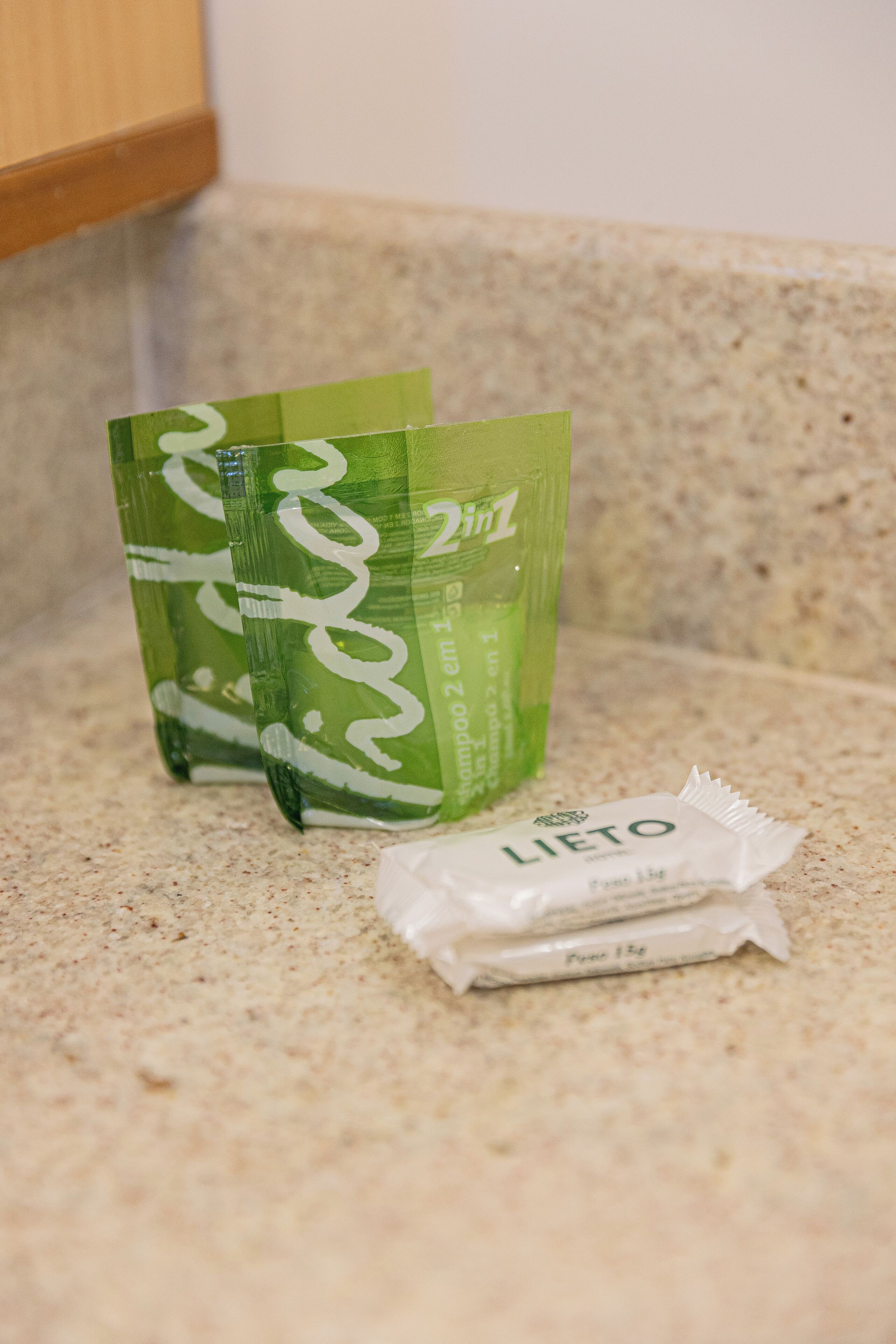 A bathroom countertop with a green bottle of shampoo, a green bottle of conditioner, and a packet of body wash.