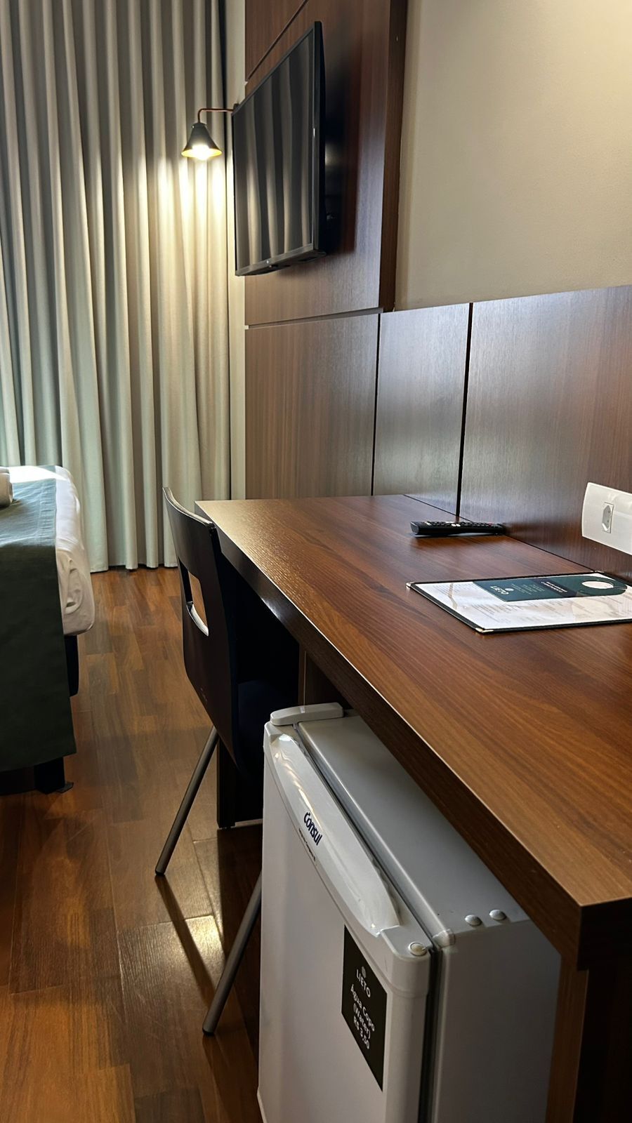 Ein kompaktes Hotelzimmer mit Schreibtisch aus Holz, einem kleinen Kühlschrank, einem flachbildfernseher an der Wand und einem Fenster mit vertikalen Jalousien.