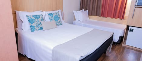 Cama doble con cabecera de madera, ropa de cama blanca y almohadas decorativas. Se ve una mesita de noche y un refrigerador.