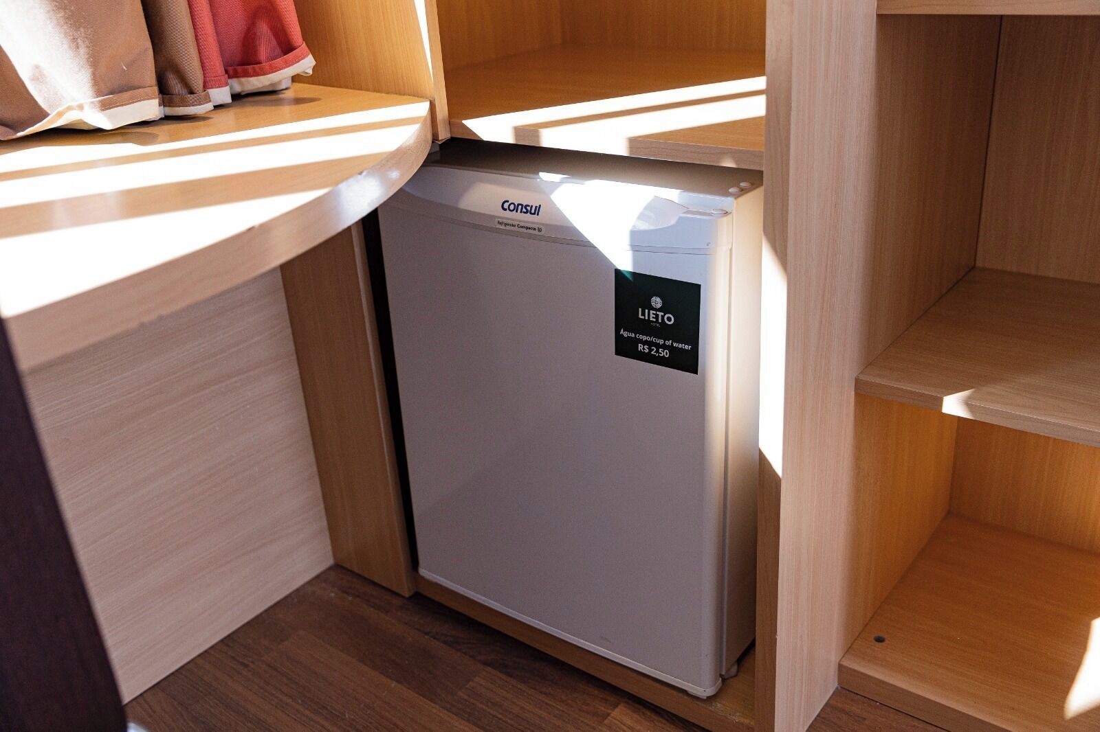 Un lavavajillas compacto y blanco con etiqueta LIETO de ahorro de agua, ubicado en un rincón de una cocina de madera con encimera de madera y un pequeño estante superior.