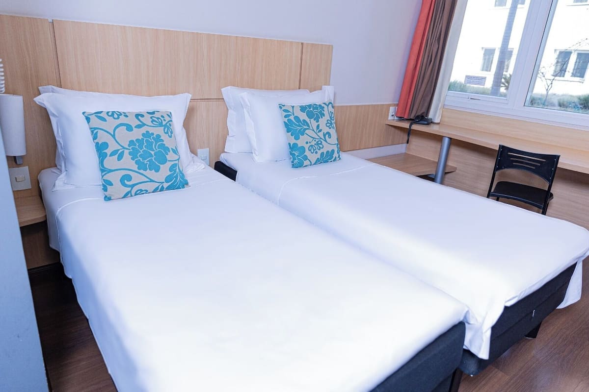 Ein Hotelzimmer mit zwei Einzelbetten, einem Holzkopfteil, einem Fenster mit Vorhängen und einem Sessel.