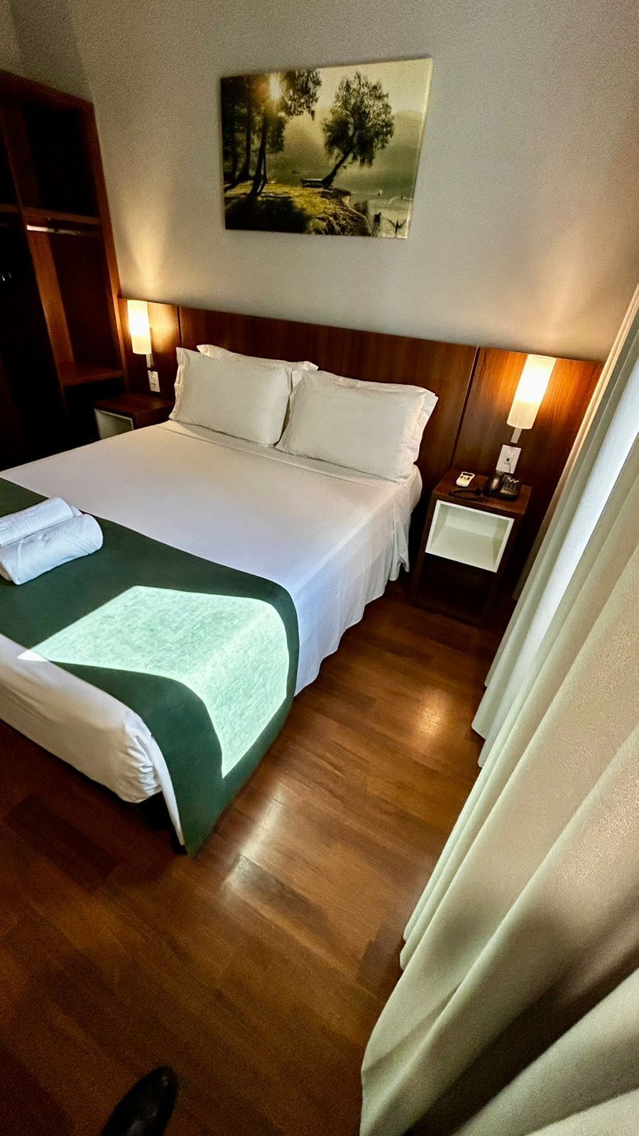 Quarto de hotel com uma cama grande, uma mesa de cabeceira com abajur, um quadro na parede e piso de madeira.