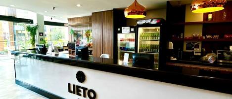 Zona de recepción de un hotel, con mostrador, un cartel que dice 'LIETO HOTEL' y un refrigerador lleno de bebidas.