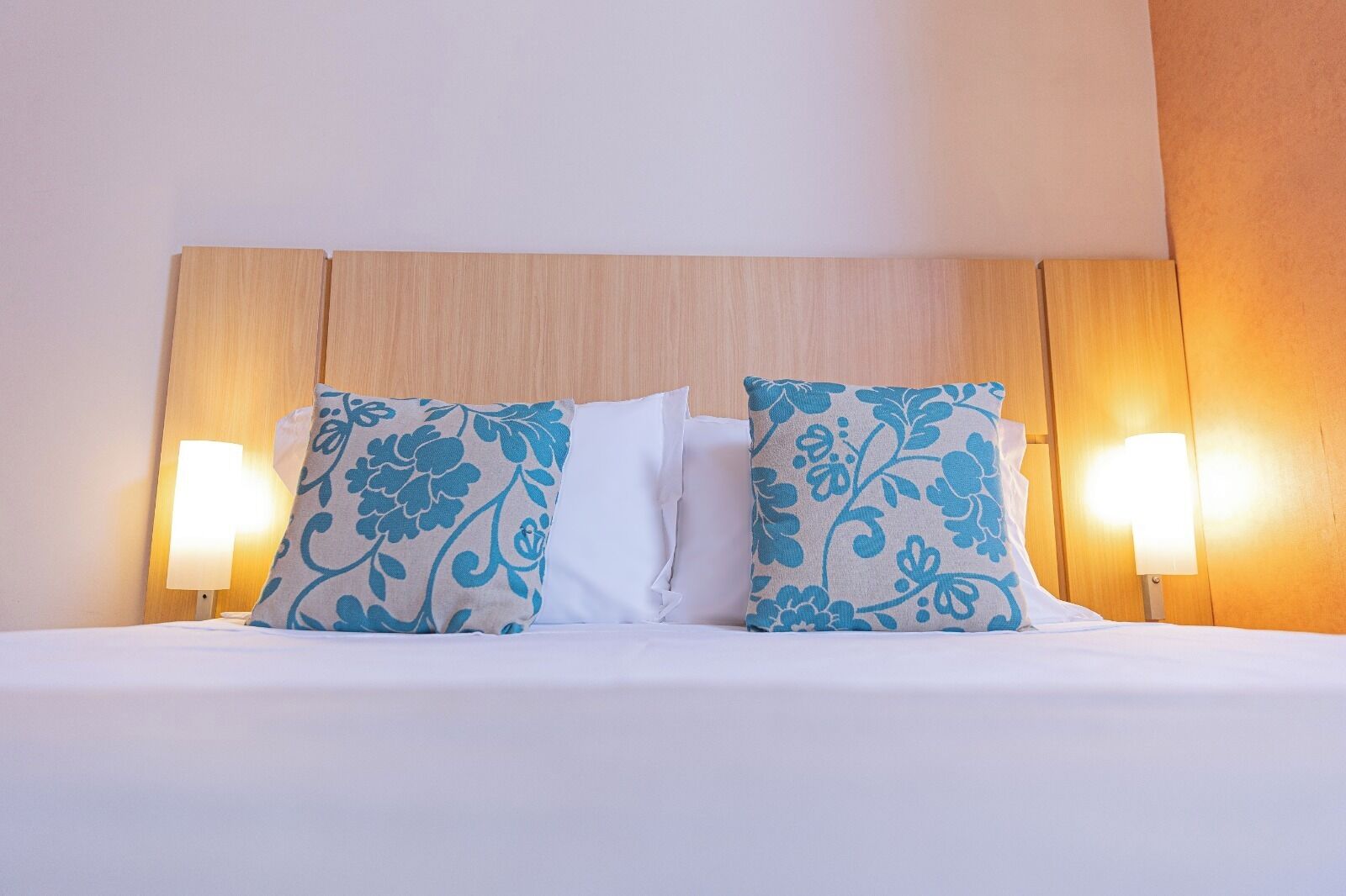 A hotel room with a wooden headboard, two decorative pillows with a floral pattern, and two wall-mounted lamps.