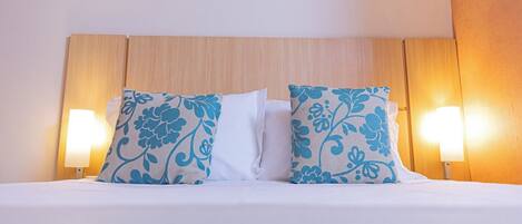 Una habitación de hotel con cabecera de madera, dos almohadones decorativos con estampado floral y dos lámparas fijadas a la pared.