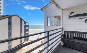 Ein Balkon mit Blick auf Strand und Meer, eine hölzerne Bank und eine Delfin-Skulptur.
