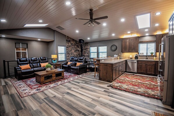 A modern living room with a leather sofa, a coffee table, and a fireplace. The kitchen features wooden cabinets and a stainless steel refrigerator.