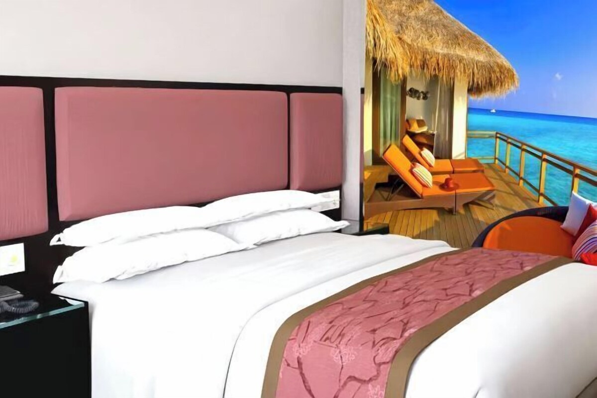 A hotel room with a bed, a nightstand, a window with a view of the ocean, and a thatched-roof balcony.
