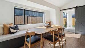 Een moderne eethoek met een houten tafel en stoelen, een bank met kussens, en een groot raam met uitzicht op een houten schutting.