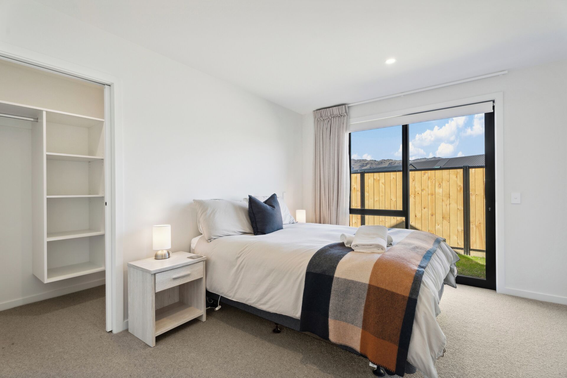 A bedroom with a bed, a nightstand, a wardrobe, and a large window with a view of a wooden fence and hills.