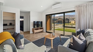 Een moderne woonkamer met een flatscreen televisie, een houten salontafel en een bank met kussens. Er is een schuifdeur naar een balkon met uitzicht op huizen en heuvels.