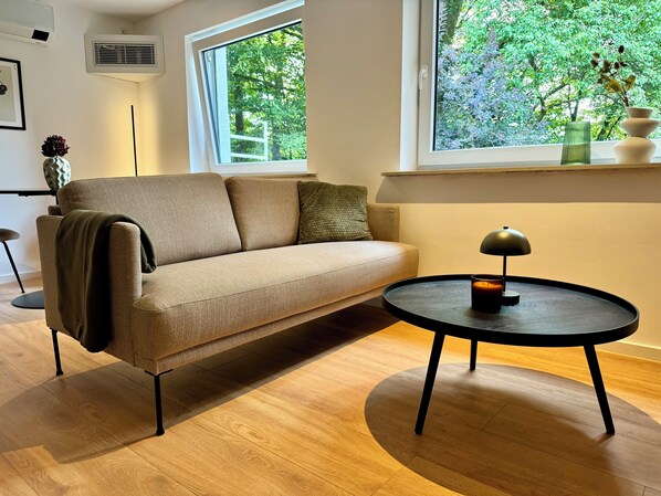 Un salon moderne avec un canapé beige, une petite table d’appoint ronde noire surmontée d’une lampe, et une fenêtre donnant sur un paysage verdoyant.
