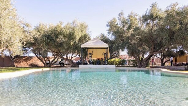 A clear swimming pool with a gazebo in the center, surrounded by trees and a building in the background. - Riad salix d'agafay (Marrakech)