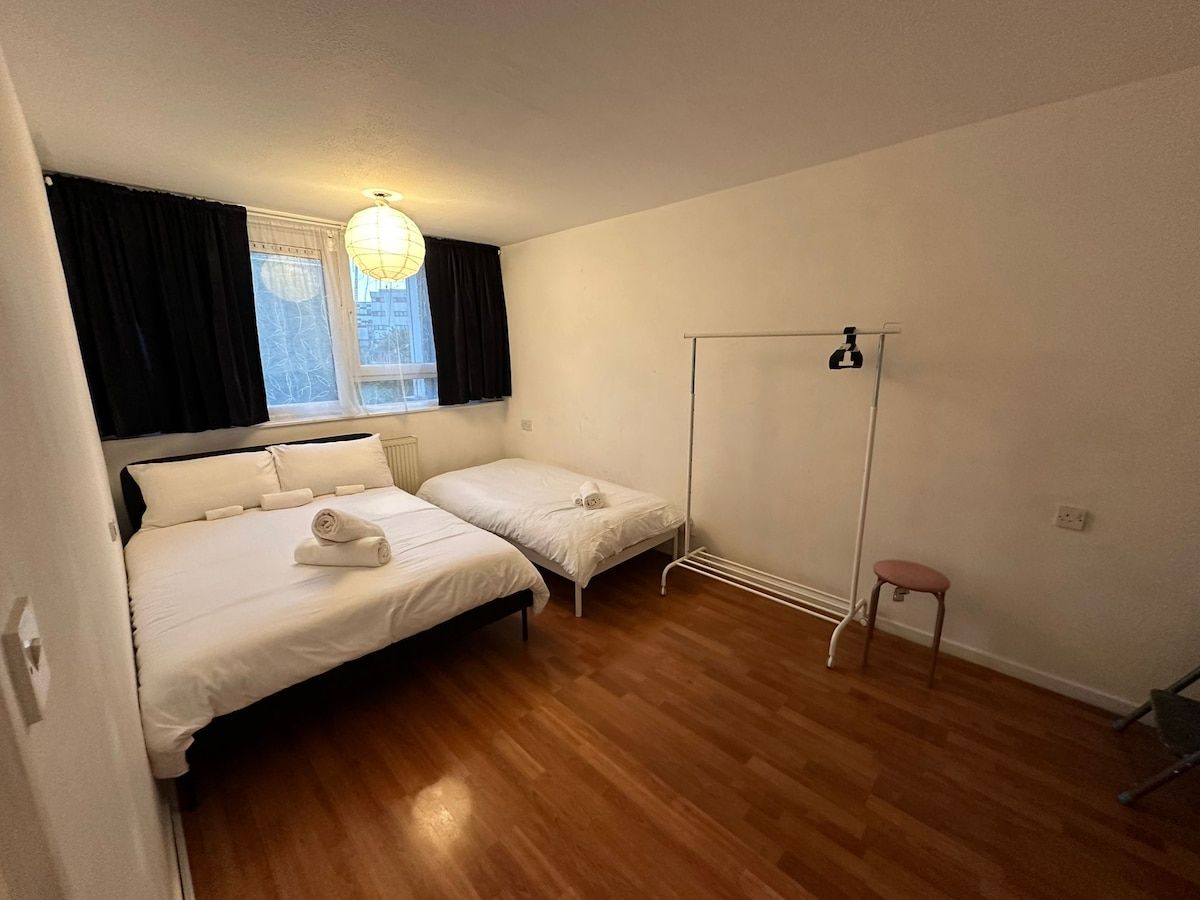 Een kamer met twee bedden, een radiator, een raam met gordijnen, een hanglamp en een klein krukje.