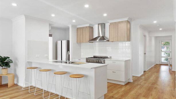 Une cuisine moderne avec des meubles blancs, un îlot central doté de tabourets, une hotte en acier inoxydable et une vue sur le salon adjacent.