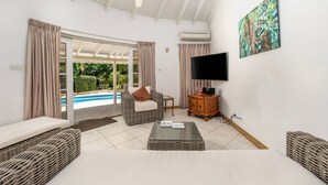 A spacious living room with a ceiling fan, a flat-screen TV, and a large window overlooking a swimming pool. - Vida Mejor--Poolside (Private Pool) (Holetown)