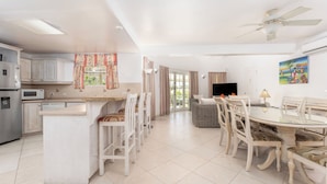 A modern kitchen with white cabinetry, a central island, and a dining area with white chairs and a glass table. - Vida Mejor West Pool (Private Pool) (Holetown)