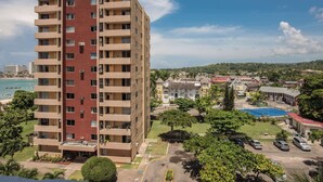 A multi-story building with balconies, a parking area, and a view of a beach and town in the background. - Emerald One Bedroom Suite (Ocho Rios)
