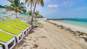 A beach with rows of green and white lounge chairs, palm trees, and a thatched-roof building. - Waterfront Colony Beach Duplex! w/ Golf Cart! (Key Colony Beach)