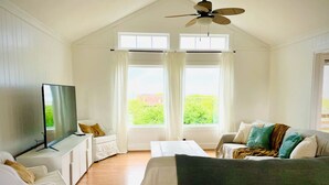 A spacious room with a large bed, a white sofa, and a TV on a stand. There are two windows with curtains, and a ceiling fan. - 3-bedroom 2 bath cottage-Walk to beach or golf
Course.Hot Tub and outside shower (Kitty Hawk)
