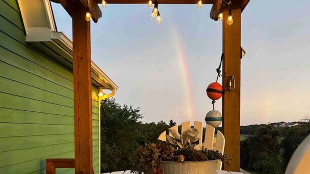 A wooden deck with a round table and white chairs, a hanging light fixture, and a rainbow in the background. - 3-bedroom 2 bath cottage-Walk to beach or golf
Course.Hot Tub and outside shower (Kitty Hawk)