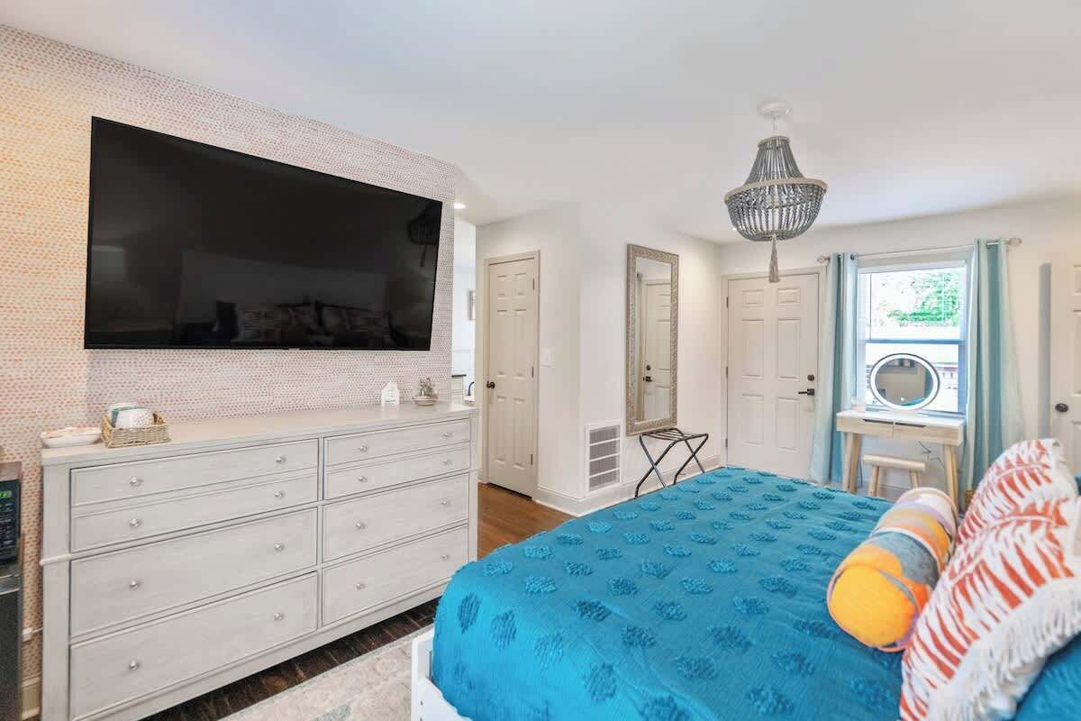 A bedroom with a large flat-screen TV, a dresser, a bed with a blue bedspread, a chandelier, and a window with curtains.