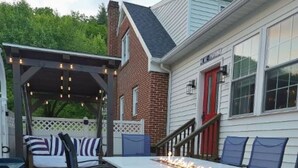 An outdoor patio with a stone table and chairs, a pergola with string lights, and a brick house in the background.