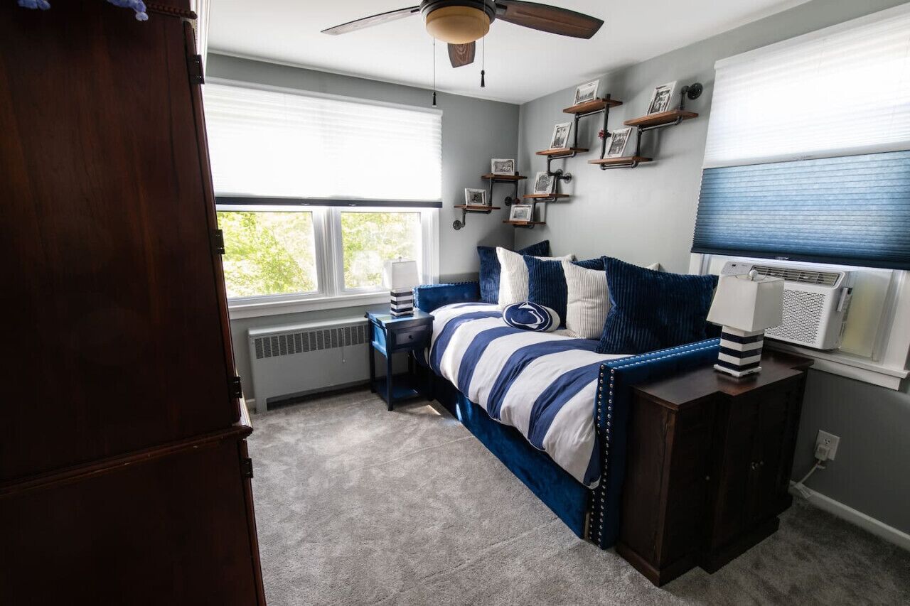 Une chambre à coucher comprenant un lit, une table de chevet, un ventilateur de plafond, une fenêtre avec des stores et une étagère murale décorée de tableaux.