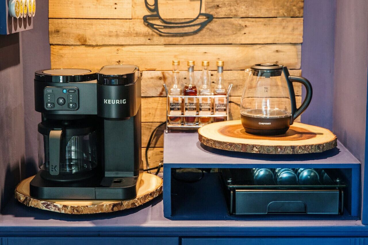 Una estación de café con una cafetera Keurig, una jarra de vidrio y una taza sobre un posavasos de madera. También hay varias botellas de sirope y un recipiente con cápsulas de café.