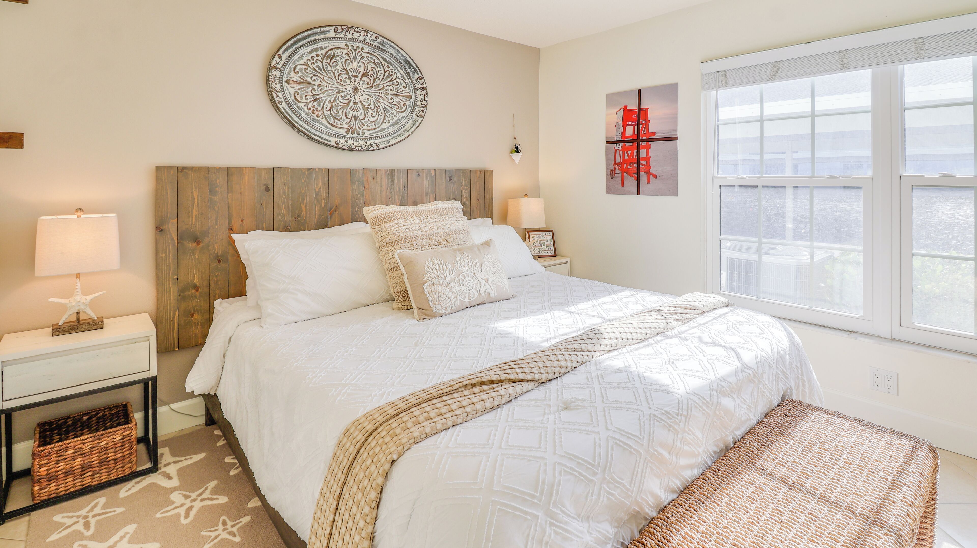 A bedroom with a wooden headboard, a large bed with white bedding, a starfish decoration on the nightstand, a woven throw blanket, and a view of the city through the window.