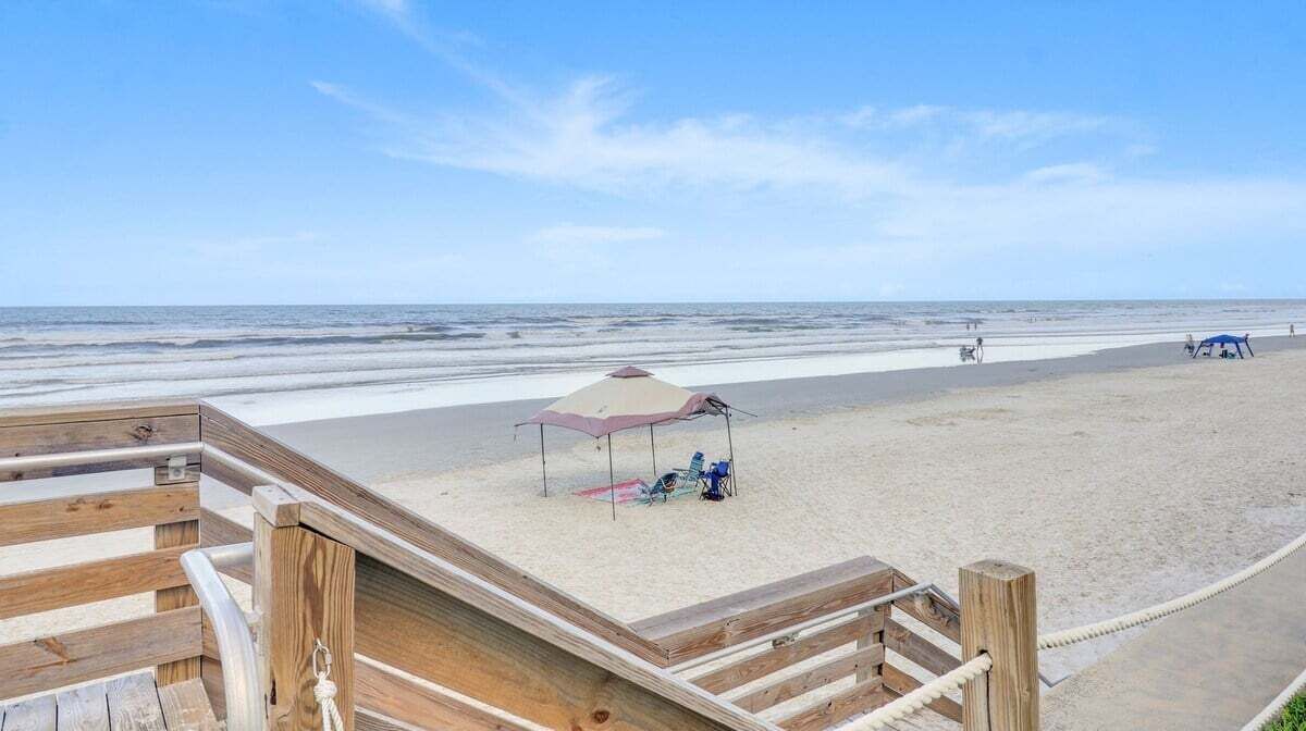 Eine Holzterrasse führt zu einem Sandstrand, mit Strandmuschel und Stühlen.