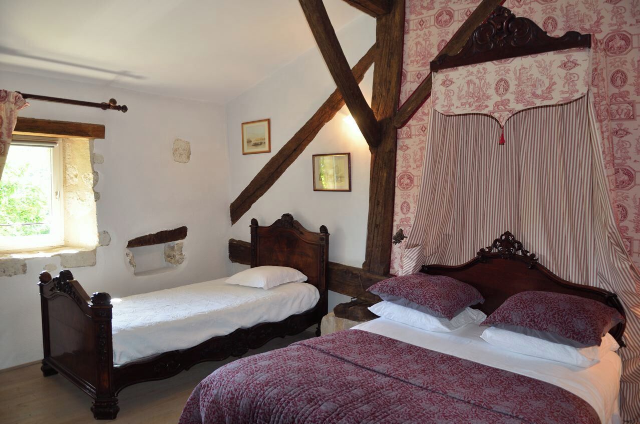 A bedroom with two wooden beds, one with a canopy and one without. There are framed pictures on the wall and a window with a curtain.