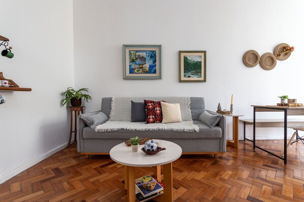 Una sala de estar moderna con un sofá gris, una mesa de centro redonda y un suelo de madera con diseño de espiga. Hay dos obras de arte enmarcadas en la pared, una planta pequeña sobre una mesita auxiliar y un cuenco decorativo en la mesa de centro.