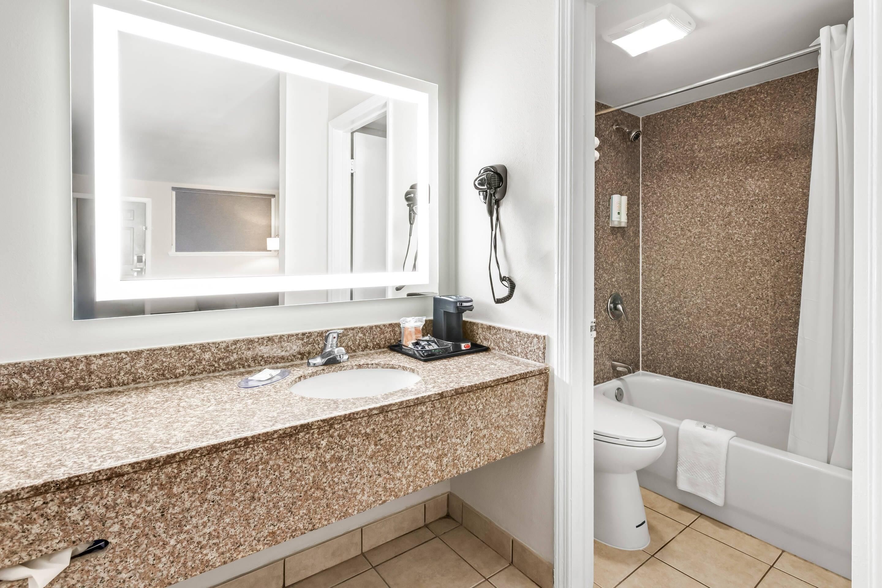 Une salle de bain avec un lavabo doté d’un plan de travail en granit, un grand miroir, un sèche-cheveux et une baignoire.