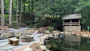 A stone patio with a fire pit, a pond, and a wooden structure with a balcony. - The Camp - Luxe Blue Ridge Condo (Burnsville)