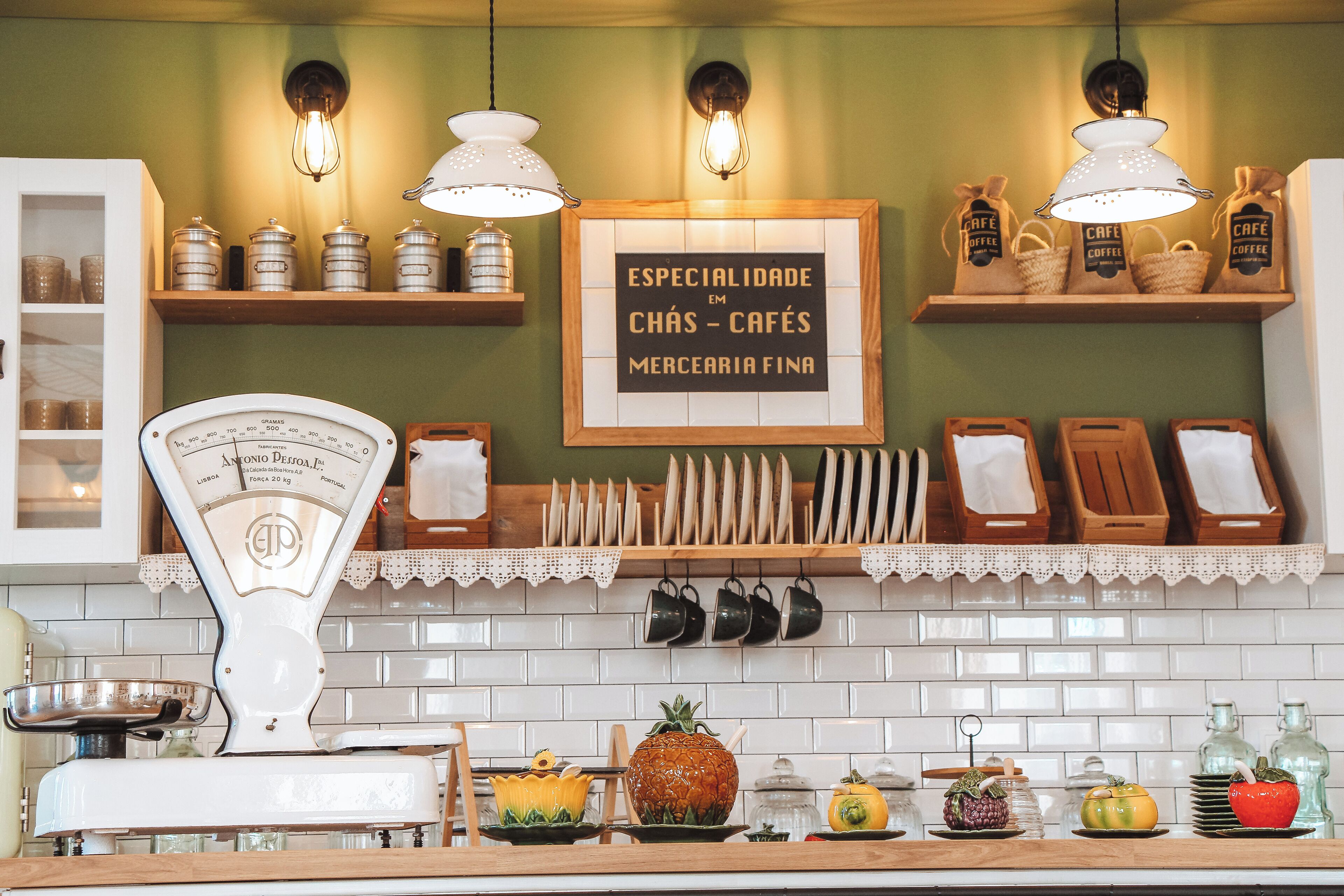 Una cocina con una balanza, tazas colgadas y un cartel que dice "Especialidade em Chás - Cafés Mercaria Fina".