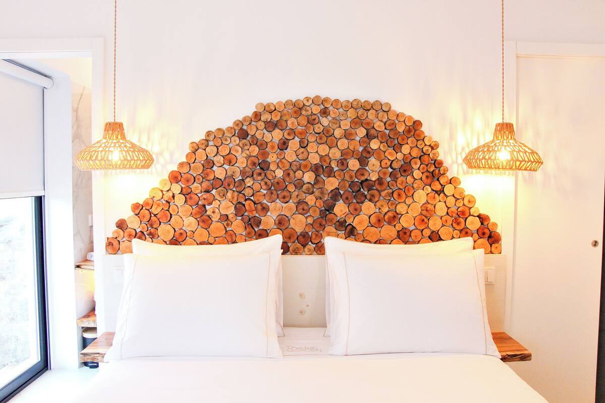 A bed with white bedding and two white pillows, a wall decorated with stacked logs, and two hanging wicker lamps.