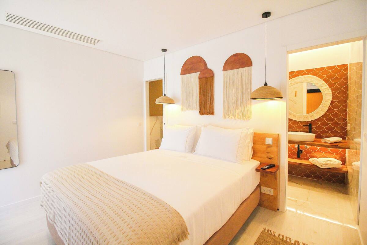 A neatly made bed with white linens, a bedside table with a remote control, and a bathroom with a mirror, sink, and towel rack.