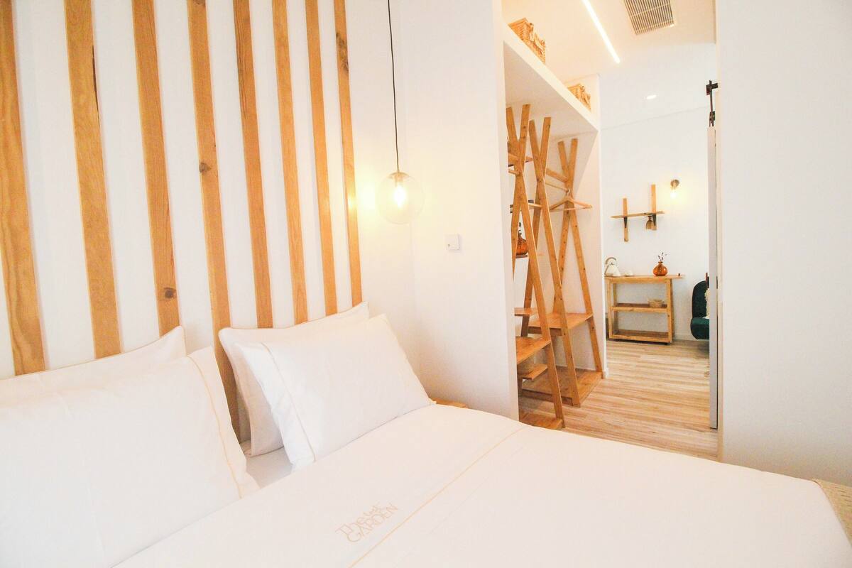 A hotel room with a bed, wooden slats on the wall, a ladder, and a small wooden table with a vase.