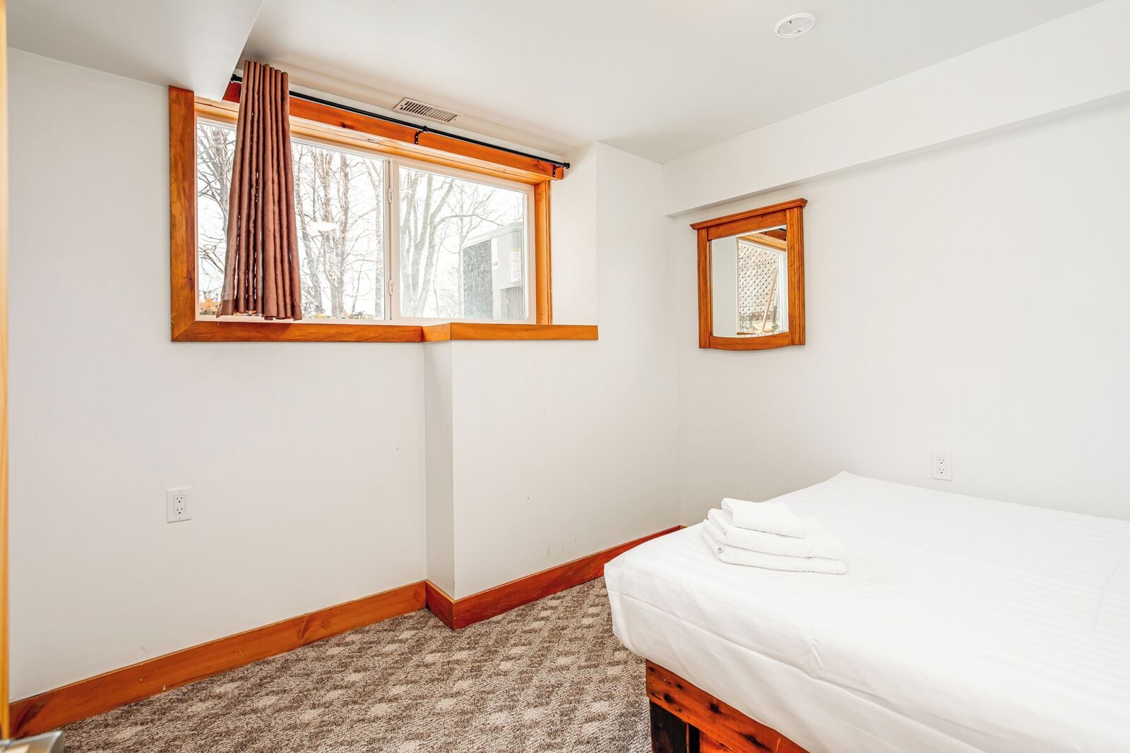 A bedroom with a single bed, a window with curtains, a small mirror, and a carpeted floor.