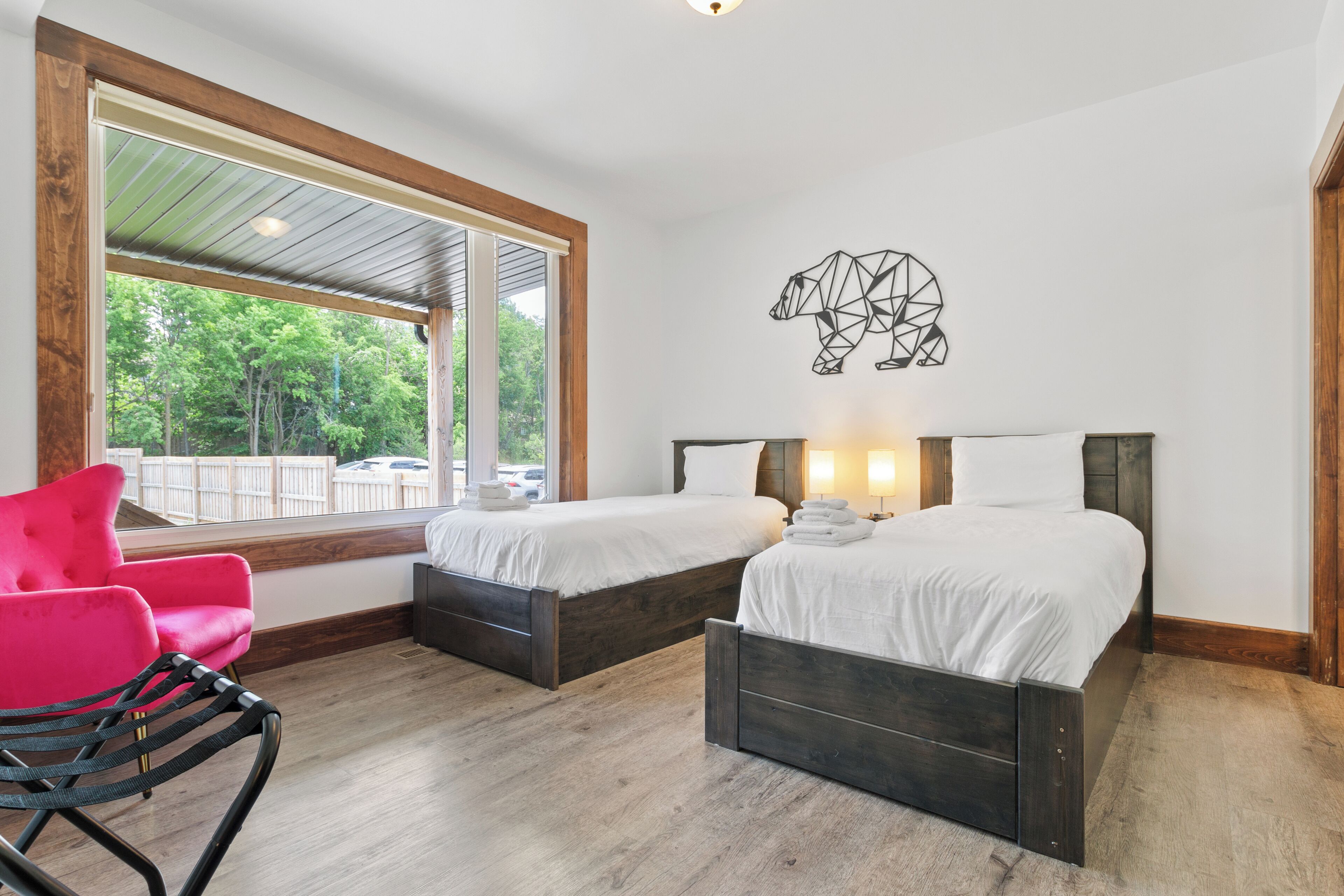 A bedroom with two beds, a large window, a red chair, and a bear wall art piece.