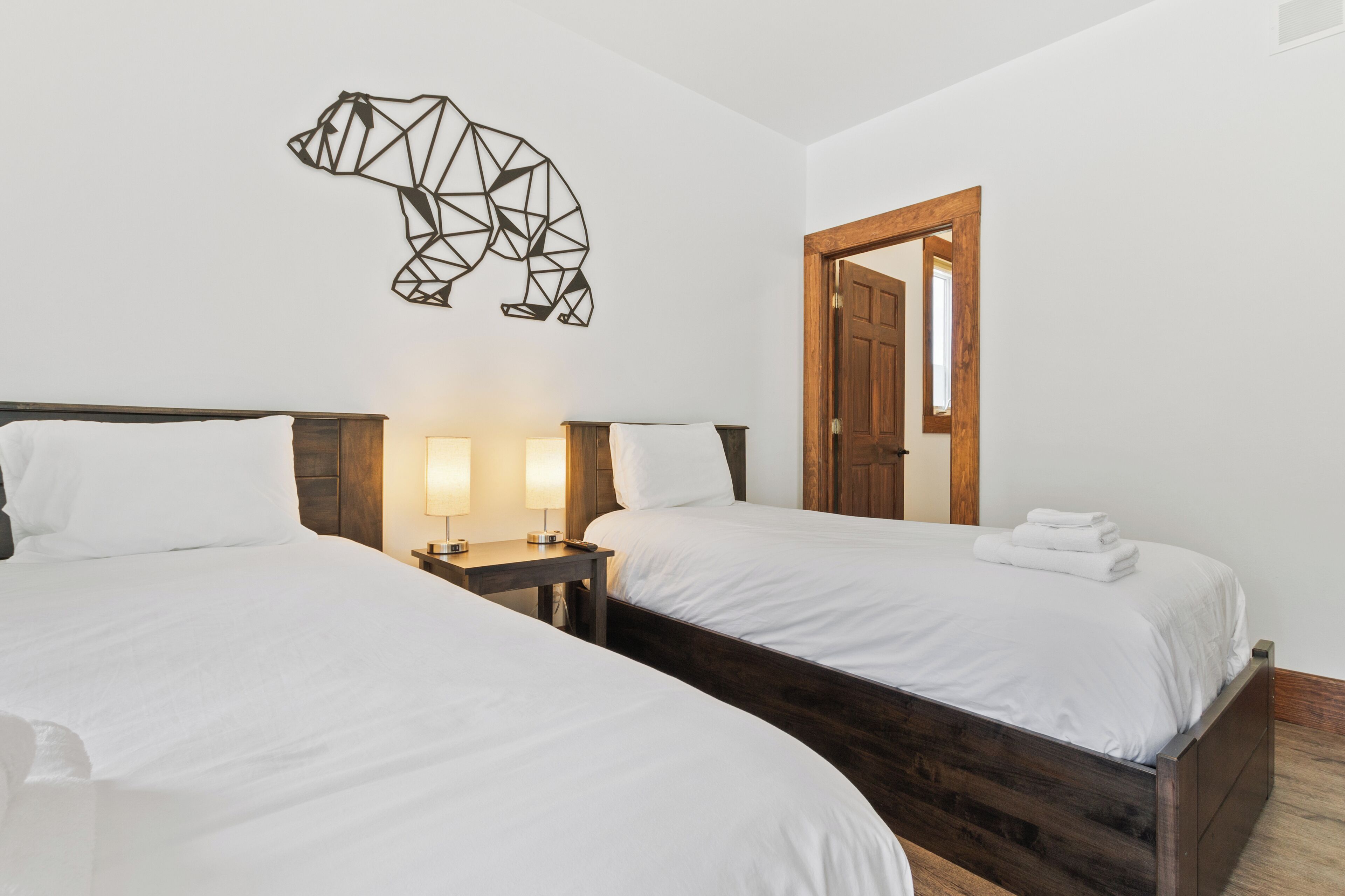 Une chambre d’hôtel avec deux lits, une tête de lit en bois, une œuvre d’art géométrique représentant un ours accrochée au mur, une table de chevet avec une lampe et une porte en bois.