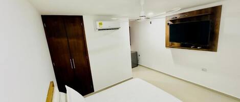 Une chambre comprenant un lit, une armoire en bois, un appareil de climatisation, un ventilateur de plafond et un téléviseur fixé au mur.